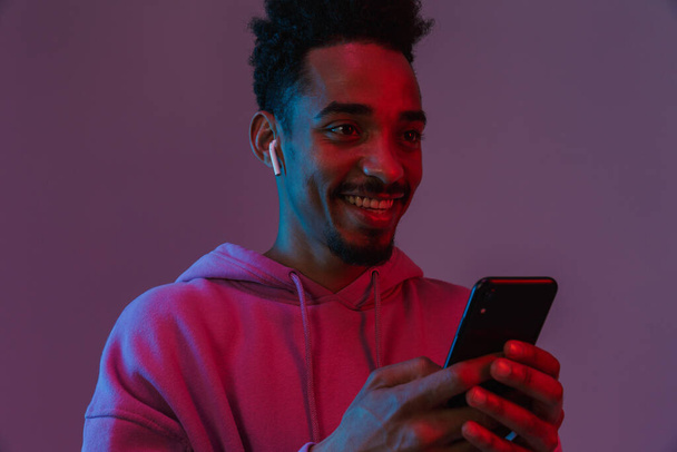 Portrait of laughing african american man in colorful hoodie talking on cellphone with earpod isolated over violet background - Valokuva, kuva