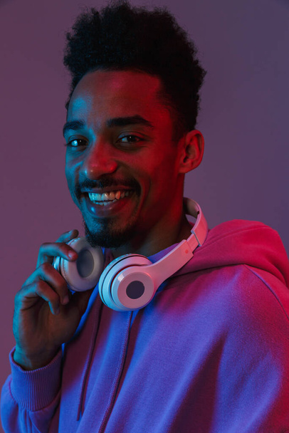 Portrait of happy african american man in colorful hoodie posing with headphones isolated over violet background - Φωτογραφία, εικόνα