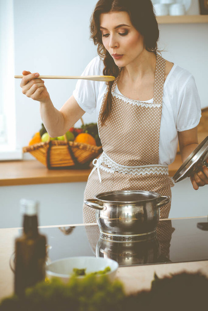 Young brunette woman cooking soup in kitchen. Housewife holding wooden spoon in her hand. Food and health concept - Fotó, kép