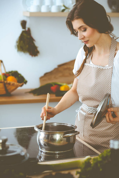Young brunette woman cooking soup in kitchen. Housewife holding wooden spoon in her hand. Food and health concept - Fotó, kép