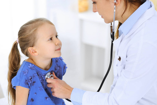 Доктор осматривает маленькую девочку стетоскопом. Счастливого улыбающегося ребенка при обычном медицинском осмотре. Медицина и здравоохранение - Фото, изображение
