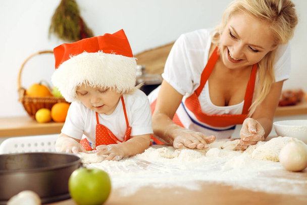 Little girl and her blonde mom in red aprons playing and laughing while kneading the dough in kitchen. Homemade pastry for bread, pizza or bake cookies. Family fun and cooking concept - Фото, изображение