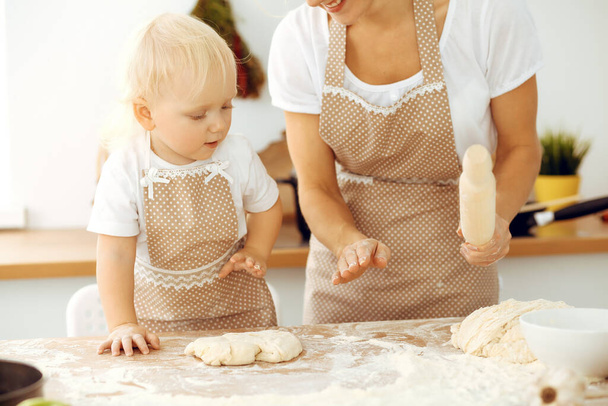 Little girl and her blonde mom in beige aprons playing and laughing while kneading the dough in kitchen. Homemade pastry for bread, pizza or bake cookies. Family fun and cooking concept - Photo, image