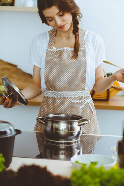 Young brunette woman cooking soup in kitchen. Housewife holding wooden spoon in her hand. Food and health concept - 写真・画像