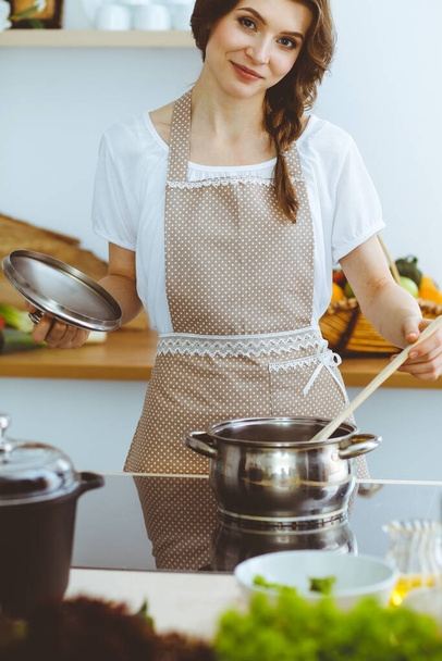 Young brunette woman cooking soup in kitchen. Housewife holding wooden spoon in her hand. Food and health concept - Foto, Imagen
