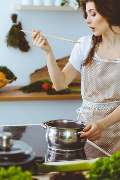 Young brunette woman cooking soup in kitchen. Housewife holding wooden spoon in her hand. Food and health concept - Foto, Imagen