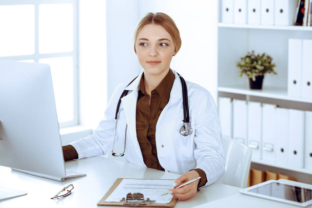 Young woman doctor at work in hospital looking at desktop pc monitor. Physician controls medication history records and exam results. Medicine and healthcare concept - Photo, image