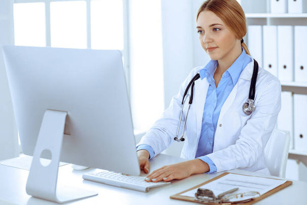 Young woman doctor at work in hospital looking at desktop pc monitor. Physician controls medication history records and exam results. Medicine and healthcare concept - Zdjęcie, obraz