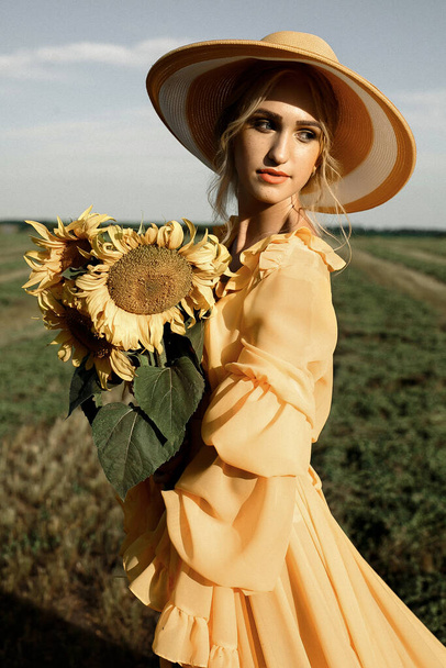 Портрет шикарной красавицы лета ярко-желтого цвета. Женщина в жёлтом платье и жёлтой шляпе держит в руках большие жёлтые подсолнухи, стоящие на зелёном поле. Летний красивый солнечный снимок. - Фото, изображение