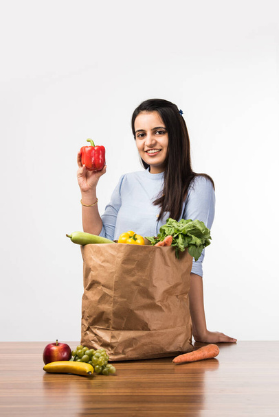 Close up portrait of a happy pretty Indian girl holding bag with vegetables and fruits standing isolated over white background - Photo, Image