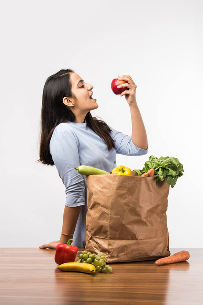 Згорніть портрет щасливої індійської дівчини, що тримає сумку з овочами й фруктами, ізольованої на білому фоні. - Фото, зображення