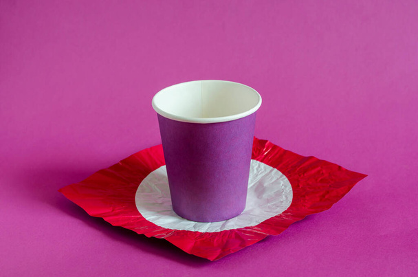 Fondo creativo con taza de papel lila y papel de color. Taza de papel para café en un soporte de papel de aluminio rojo con un círculo blanco. Vista lateral en ángulo. Enfoque selectivo
. - Foto, Imagen