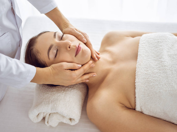 Beautiful brunette woman enjoying facial massage with closed eyes. Relaxing treatment in medicine and spa center concepts - Photo, image