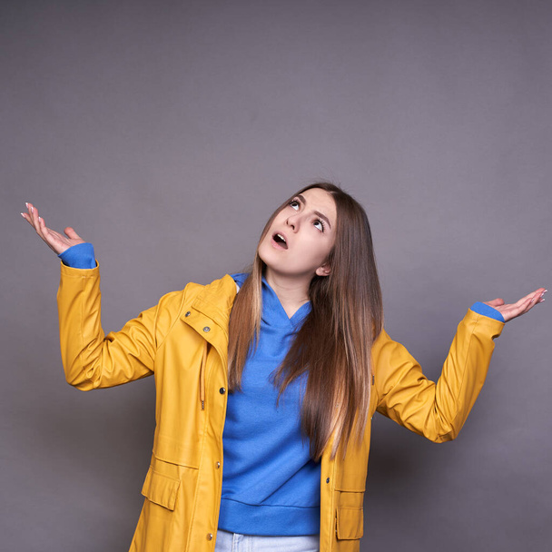 An attractive young Caucasian girl, with long blond hair, dressed in a blue sweatshirt and yellow raincoat, spread her arms to the side, looks up, it will rain soon, it seems the rain begins. - Foto, Bild