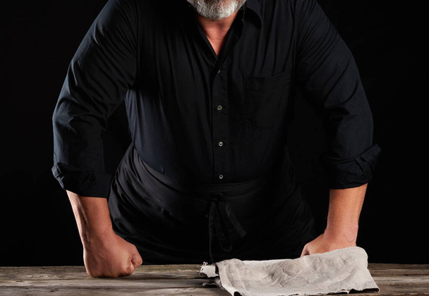 chef-kok in zwart uniform gebogen over een houten tafel en rustte zijn vuisten, zwarte achtergrond - Foto, afbeelding