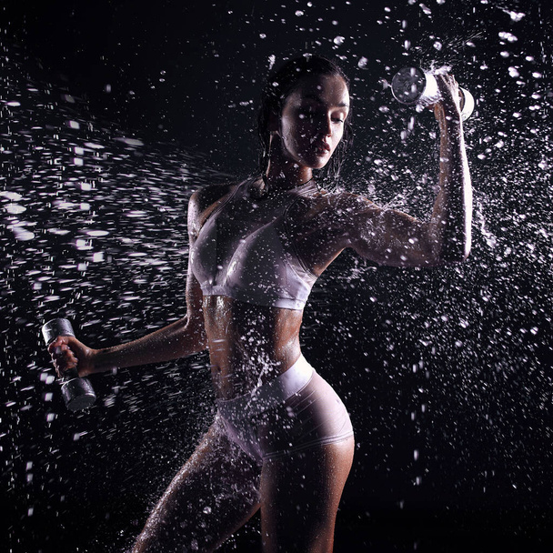 athletic woman pumping up muscles with dumbbells Beautiful athletic caucasian woman lifts dumbbells in aquastudia against a background of splashing water. Freshness sport motivation - Фото, зображення