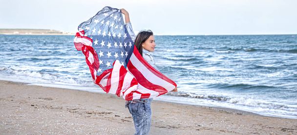 Una giovane donna con una bandiera americana corre vicino al mare. Il concetto di patriottismo e celebrazioni del giorno dell'indipendenza. - Foto, immagini