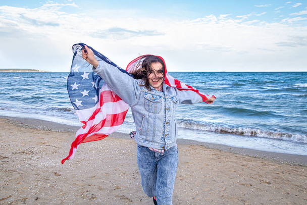 Młoda kobieta z amerykańską flagą biegnie nad morzem. Koncepcja patriotyzmu i obchodów Dnia Niepodległości. - Zdjęcie, obraz
