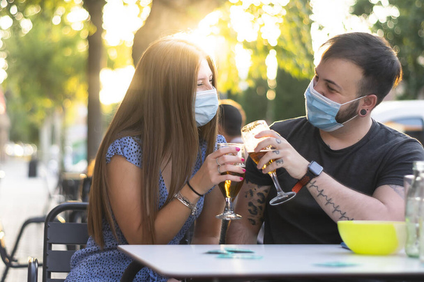Spain Madrid. Caucasian couple toasting with beer wearing sanitary mask. Reopen pubs and club after quarentine coronavirus Covid-19. Man and woman drinking beer. Cheers. People smiling wearing mask - Fotó, kép