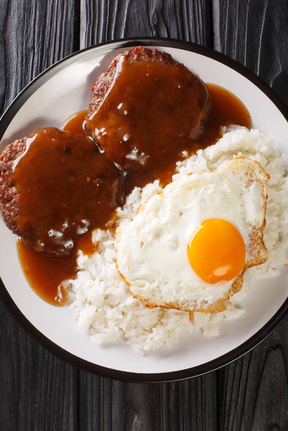 Loco moco - блюдо современной гавайской кухни, состоящее из белого риса, увенчанного гамбургером, жареным яйцом и коричневым соусом крупным планом на тарелке на столе. Вертикальный вид сверху с бова - Фото, изображение