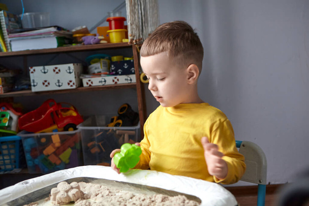 The boy is playing in his room. Educational game. A child plays kinetic sand in quarantine. Development of fine motor skills in children - Photo, Image