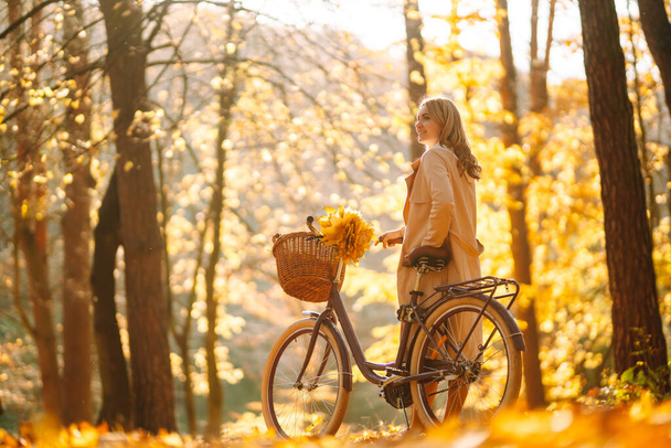 Young smiling girl with a bicycle walks in the autumn forest at sunset. - Φωτογραφία, εικόνα
