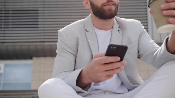 Милый бизнесмен с бородой сидит на лестнице и смотрит на финансовый отчет компании в своем смартфоне на открытом воздухе. Положительные эмоции, хорошее настроение, счастье. - Кадры, видео