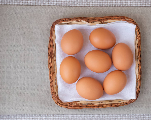 Braune frische rohe Eier liegen auf einer weißen Serviette in einem kleinen flachen Korb. Der Korb liegt auf dem Tisch vor beigem Hintergrund. Flache Lage Top wiev Kopierraum Platz für Text - Foto, Bild