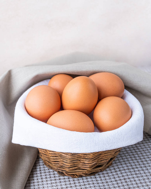 Os ovos crus frescos marrons estão em um guardanapo branco em uma pequena cesta rústica áspera. O cesto está sobre a mesa em um fundo bege. Espaço para cópia Lugar para texto Vertical
 - Foto, Imagem