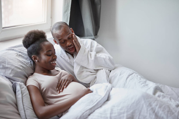 happy afro man suprisingly looks at pregnant wifes belly - Valokuva, kuva