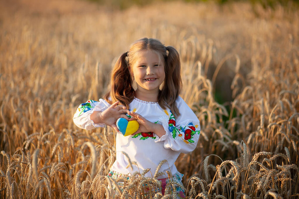 Menina vestida com uma camisa bordada (vyshyvanka) com um coração amarelo e azul em suas mãos em um campo de trigo ao pôr do sol. Dia da Independência da Ucrânia. Pequeno patriota. - Foto, Imagem