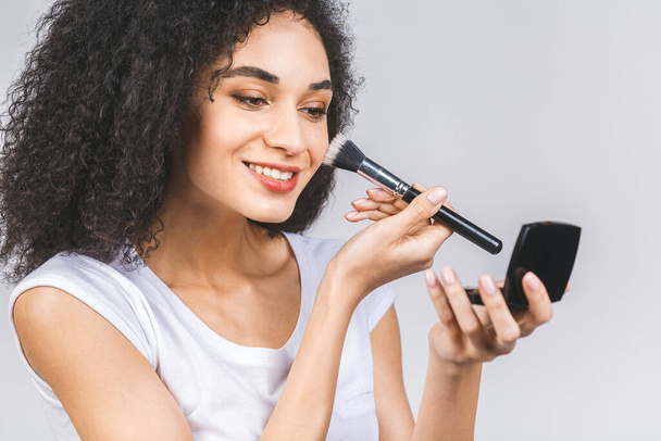 glimlachende afro-Amerikaanse vrouw aanbrengen make-up poeder of stichting met borstel geïsoleerd op een grijze achtergrond.  - Foto, afbeelding