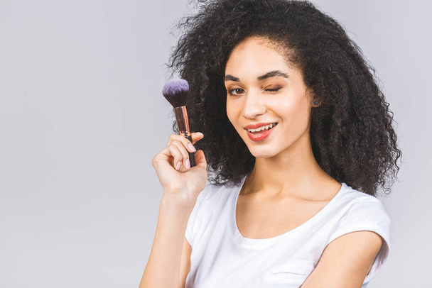 Mujer afroamericana sonriente aplicando maquillaje en polvo con cepillo aislado sobre un fondo gris
.  - Foto, imagen