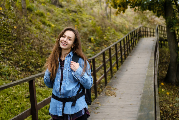 Woman tourist with a backpack walks on a bridge in the autumn forest. - Photo, Image