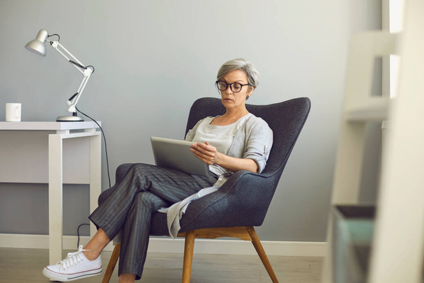 Online work education communication. Senior woman with glasses uses tablet at home. - Fotó, kép