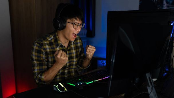 Emocionado hombre asiático pro gamer sentado a la mesa, jugando y ganando en los videojuegos en línea en un ordenador y teléfono inteligente, tecnología de juegos cibernéticos o e-sport concepto de campeonato. - Foto, Imagen