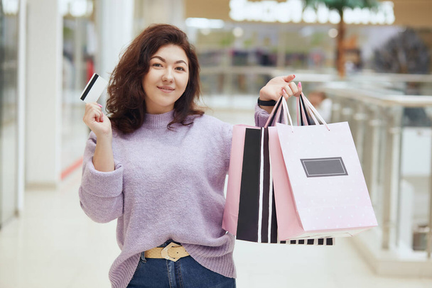 Charming lady wearing lilac sweater holding credit card and shopping bags in hands posing in shopping mall, standing with her purchases, doing shopping, pays with credit card. - Фото, изображение