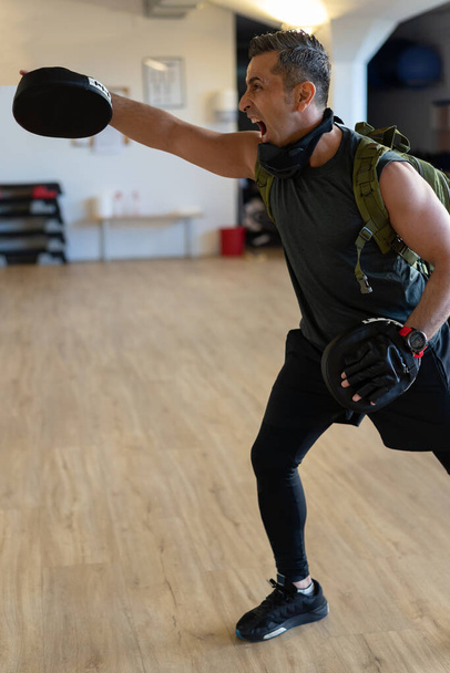 Motivated boot camp instructor stands with punch mitts in gym hall. Training with extended arm on wooden floor. Portrait for fitness concept. - Photo, Image