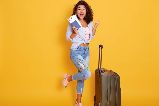 Funny happy girl posing on one leg near suitcase, holding passport with flight tickets, isolated over yellow background, clenching fists and looks happy. - Foto, Imagem