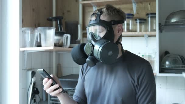 νευρικός άνθρωπος με μάσκα αερίου μιλάει smartphone στην κουζίνα στο σπίτι. προστασία από ιούς - Πλάνα, βίντεο