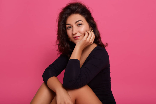 Portrait beautiful young caucasian woman wearing black shirt, sitting on floor with hands under chin isolated over pink background, looking directly at camera with confident look. - Fotoğraf, Görsel