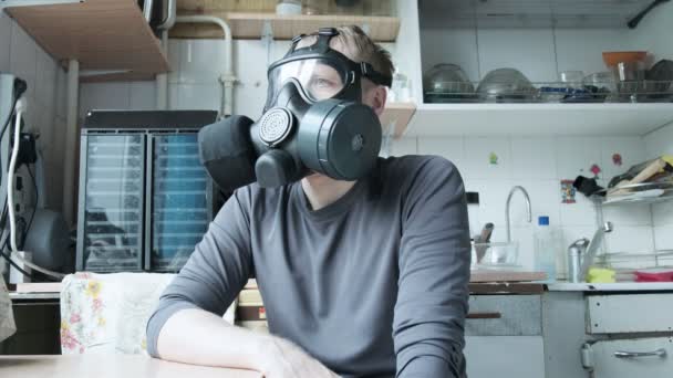 Ένας άντρας με μάσκα αερίου κάθεται στην κουζίνα στο σπίτι. χημικό όπλο, προστασία από ιούς - Πλάνα, βίντεο