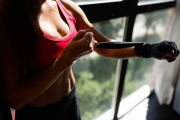 Close-up of the chest of a young woman boxer in a sports top, who ties her arm with black elastic bandages against the background of a panoramic window. - Foto, imagen