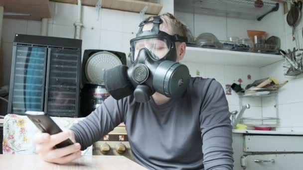 νεαρός άνδρας με μάσκα αερίου μιλάει smartphone στην κουζίνα στο σπίτι. προστασία από ιούς - Πλάνα, βίντεο