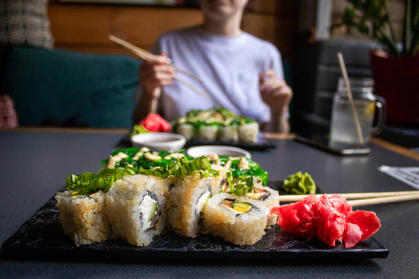 Mão de perto segurando pauzinhos de bambu com camarão nigiri enquanto mergulhando em molho de soja. Detalhe do sushi colocado na bandeja de madeira no restaurante, enquanto mão mergulho nigiri em molho de soja. Conceito de cozinha japonesa
. - Foto, Imagem