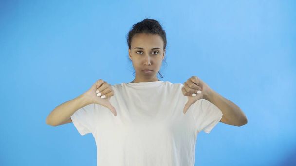 Menina bonita em uma camiseta branca mostra polegar para baixo com ambas as mãos no fundo azul
 - Foto, Imagem