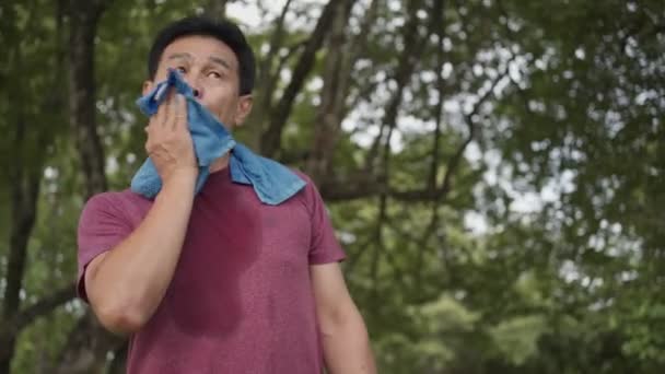 Azjatycki mężczyzna w średnim wieku wycierający pot na twarzy i szyi podczas ćwiczeń w parku, stojący pod drzewami i odpoczywający po treningu, zmęczone odnowy ciała, stan zdrowia, niski kąt - Materiał filmowy, wideo