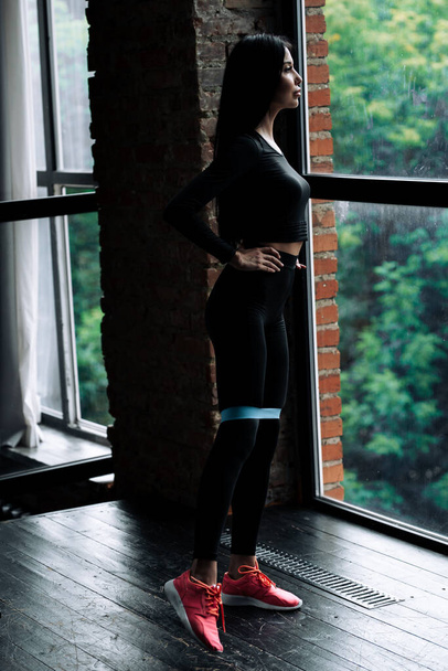 Full-length portrait of a girl who trains leg muscles using an elastic rubber expander standing in front of a panoramic window in a loft - Foto, imagen