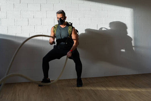 Sportsman at rope workout with training mask. Indoor on oak floor with sun and shadow. For boot camp concept. - Photo, Image