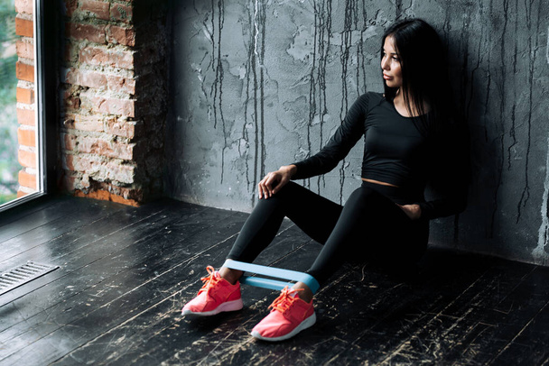Young woman sits on the floor near the wall with ankle elastic rubber expander to train leg muscles - Fotoğraf, Görsel